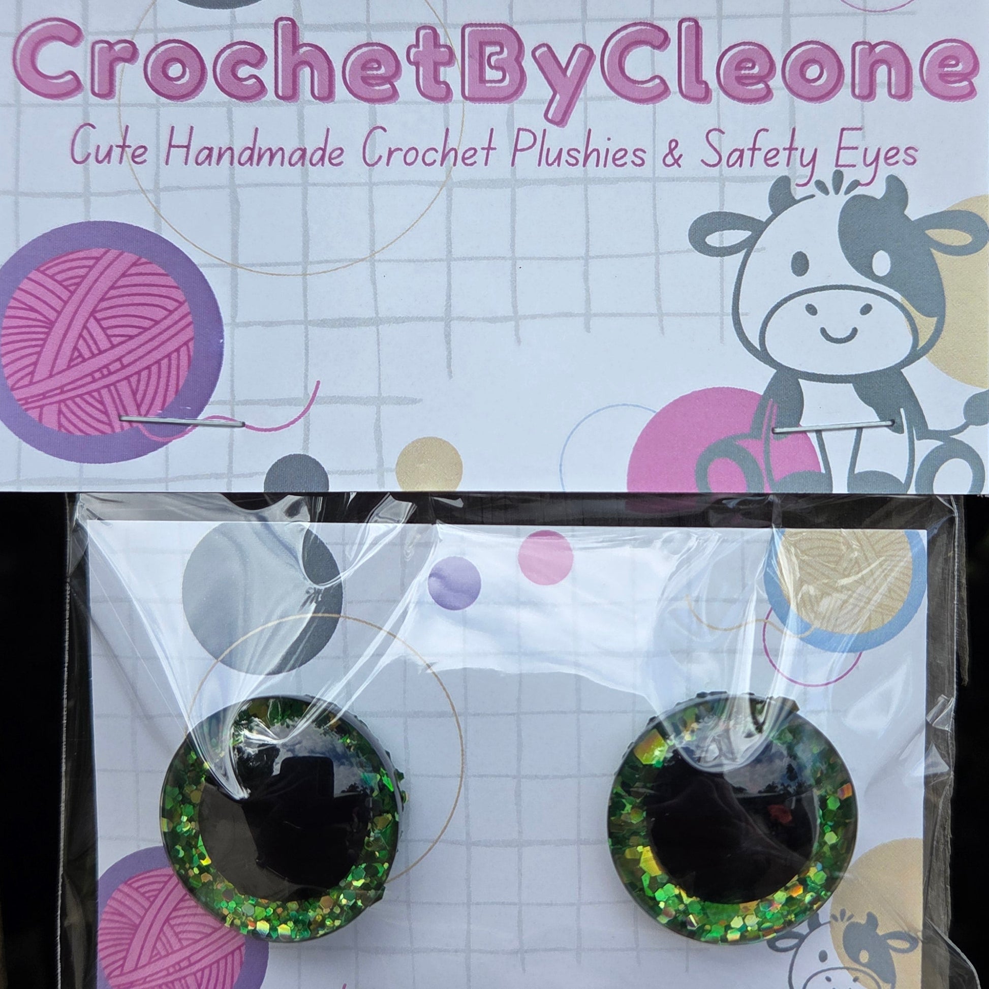 A pair of handmade crochet plush eyes in glittery green and black, packaged on a white card with colorful yarn ball graphics. The card is branded 'CrochetByCleone' and placed next to its packaging on a rustic wooden surface.