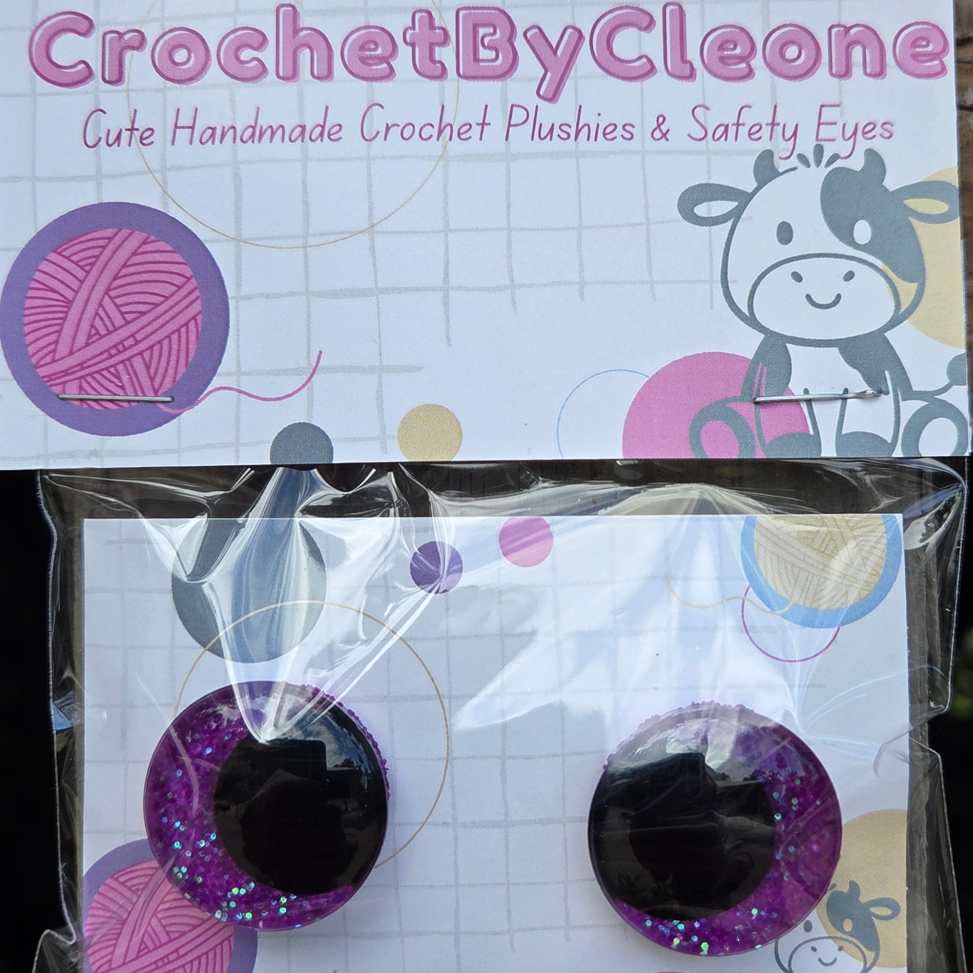 A pair of handmade crochet plastic and safety eyes in a clear plastic bag, displayed next to its branded cardboard backing on a rustic wooden surface. The brand 'CrochetByCleo' is clearly visible on the packaging.