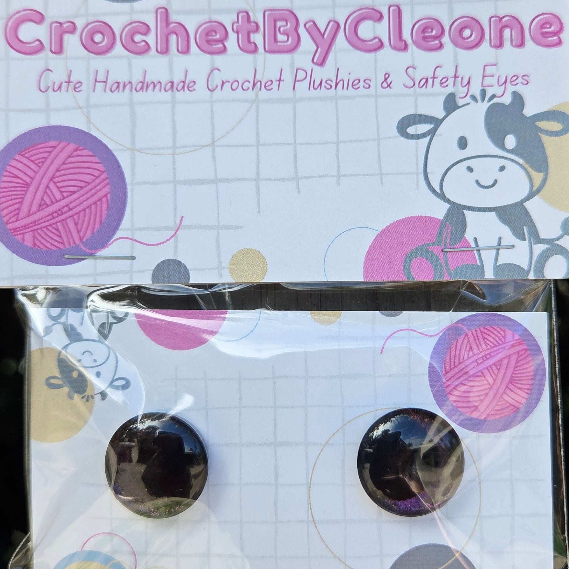 A pair of round black safety eyes with purple glitter accents, displayed on a branded backing card that reads 'CrochetByCleone' and says 'Gute Handmade Crochet Plushies & Safety Eyes.' The card has crochet-themed graphics including yarn balls, a cow, and a grid pattern, and the product is photographed in clear plastic packaging against a rustic wooden background.