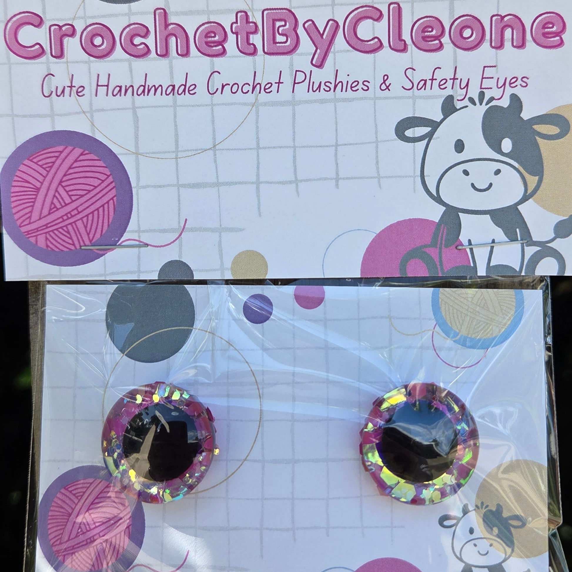 A pair of handmade crochet safety eyes in a clear plastic sleeve, displayed next to their branded packaging card. The packaging features a white background with pink and purple yarn graphics, a cartoon cow, and the brand name 'CrochetByCleo' printed vertically.