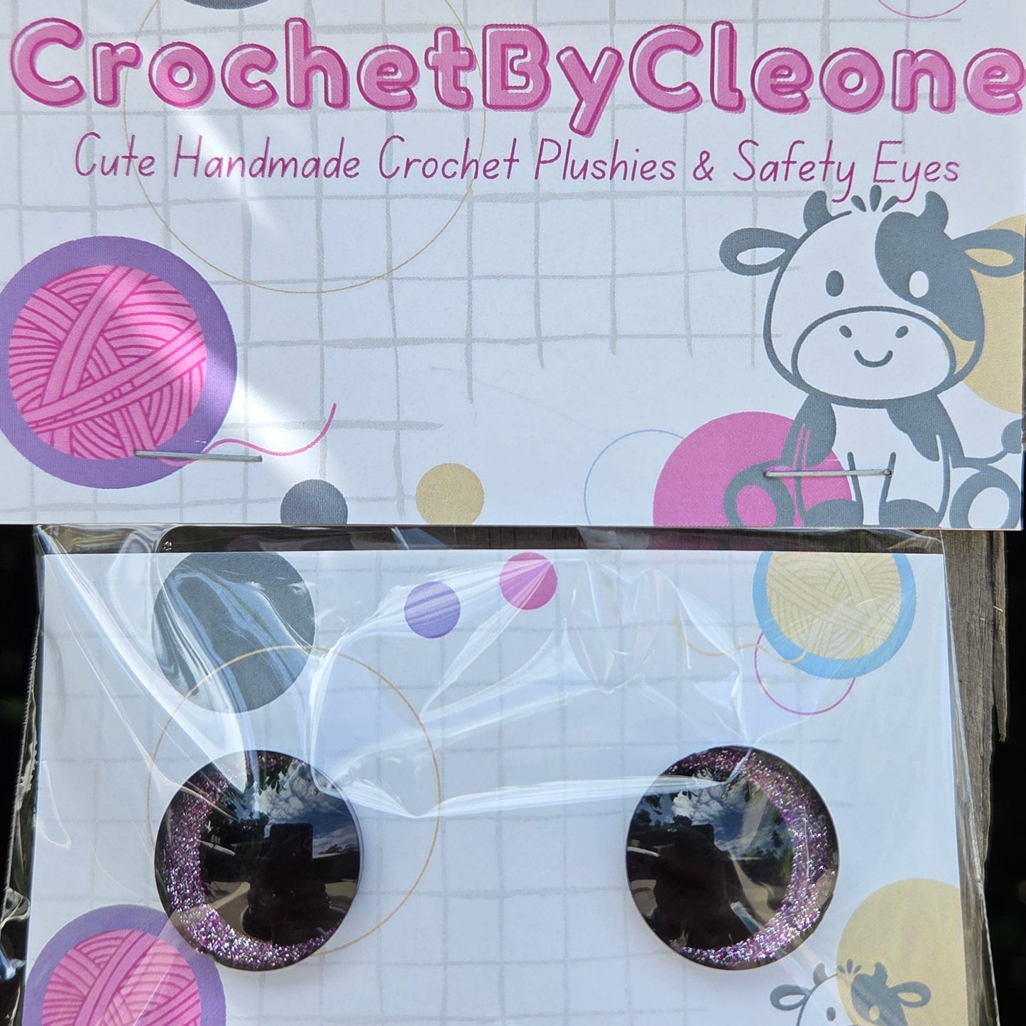 A pair of handmade crochet plushies in clear plastic packaging, displayed next to their branded cardboard backing card on a rustic wooden surface.