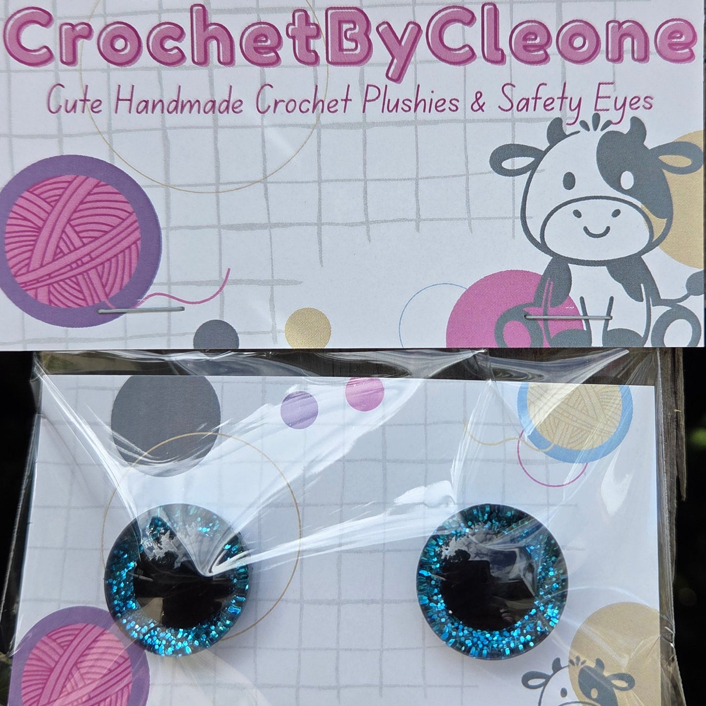 Two round glittery blue and black safety eyes packaged on a white card with crochet-themed graphics, placed on a rustic wooden surface.