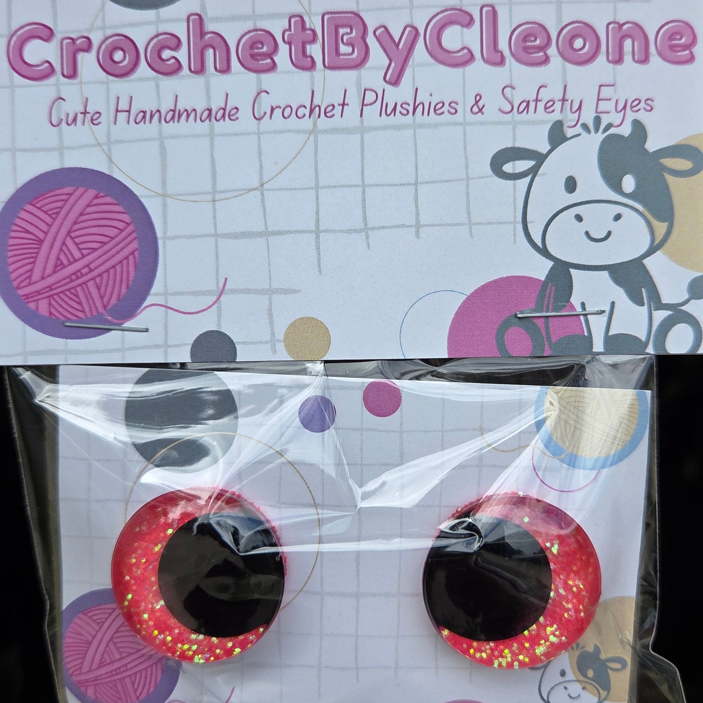 A pair of handmade crochet safety eyes by CrochetByCleo, shown with their branded packaging card and clear plastic bag on a rustic wooden surface.