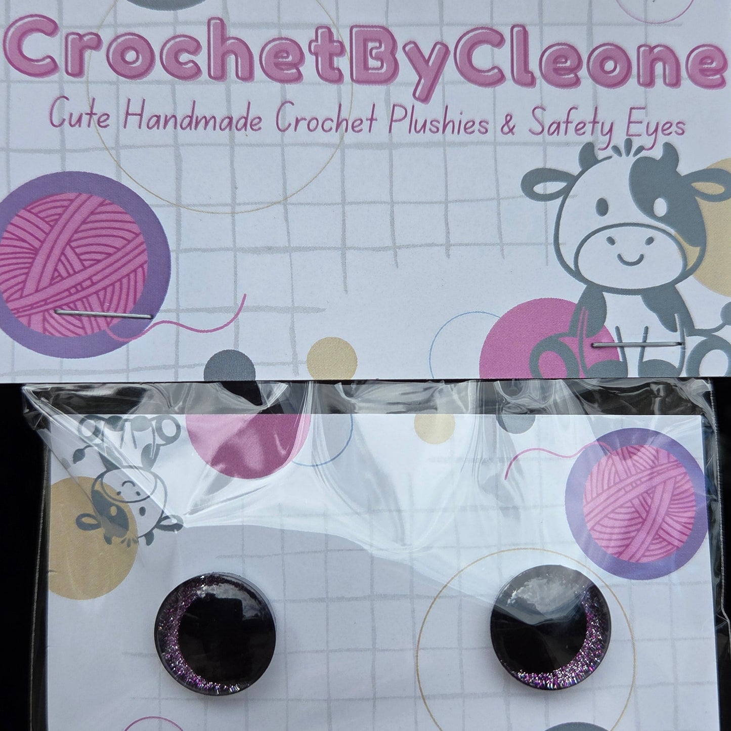 A pair of handmade crochet plushies in circular black and glittery pink eyes, packaged on a white card with crochet-themed graphics next to its branded CrochetByCleoone packaging.