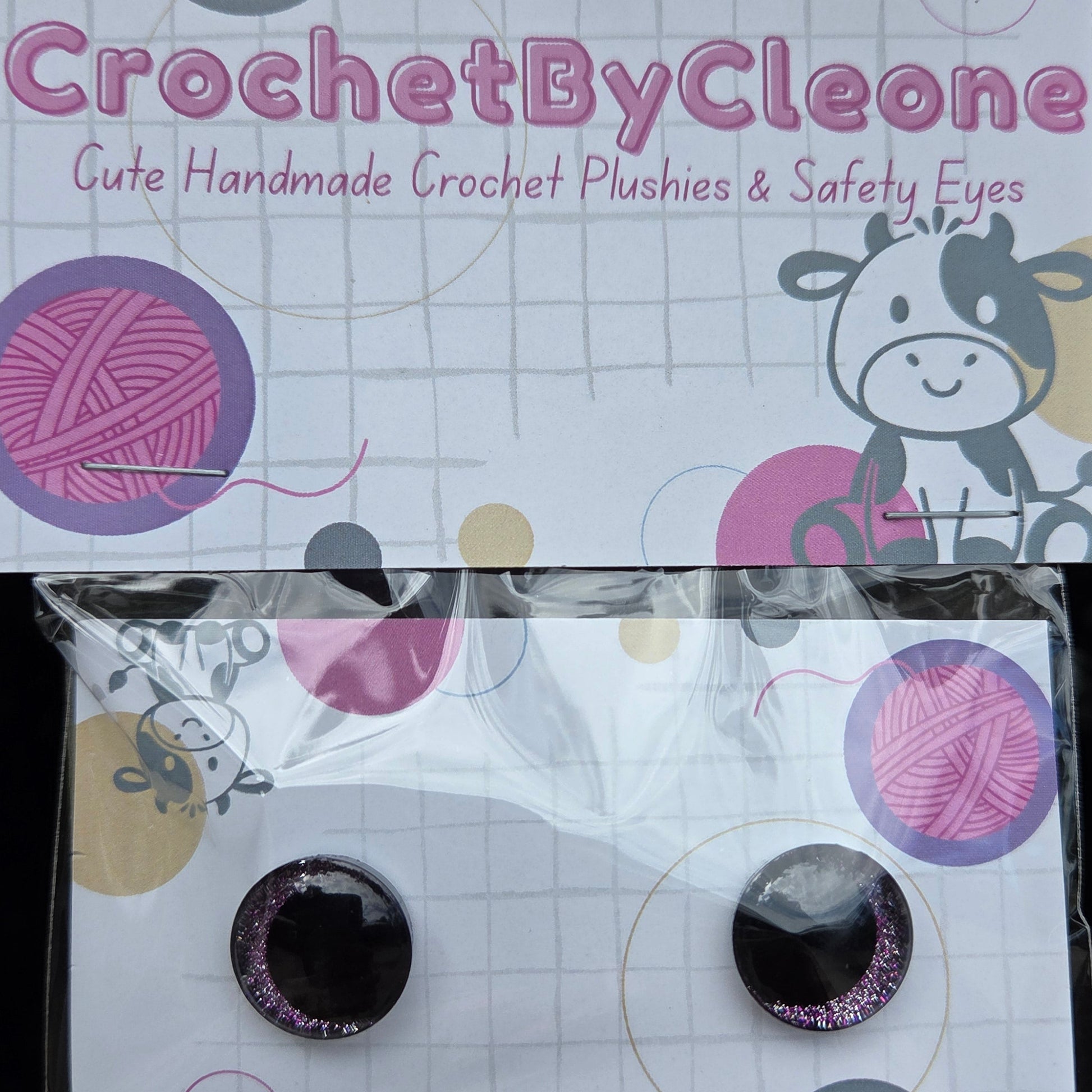 A pair of handmade crochet plushies in circular black and glittery pink eyes, packaged on a white card with crochet-themed graphics next to its branded CrochetByCleoone packaging.