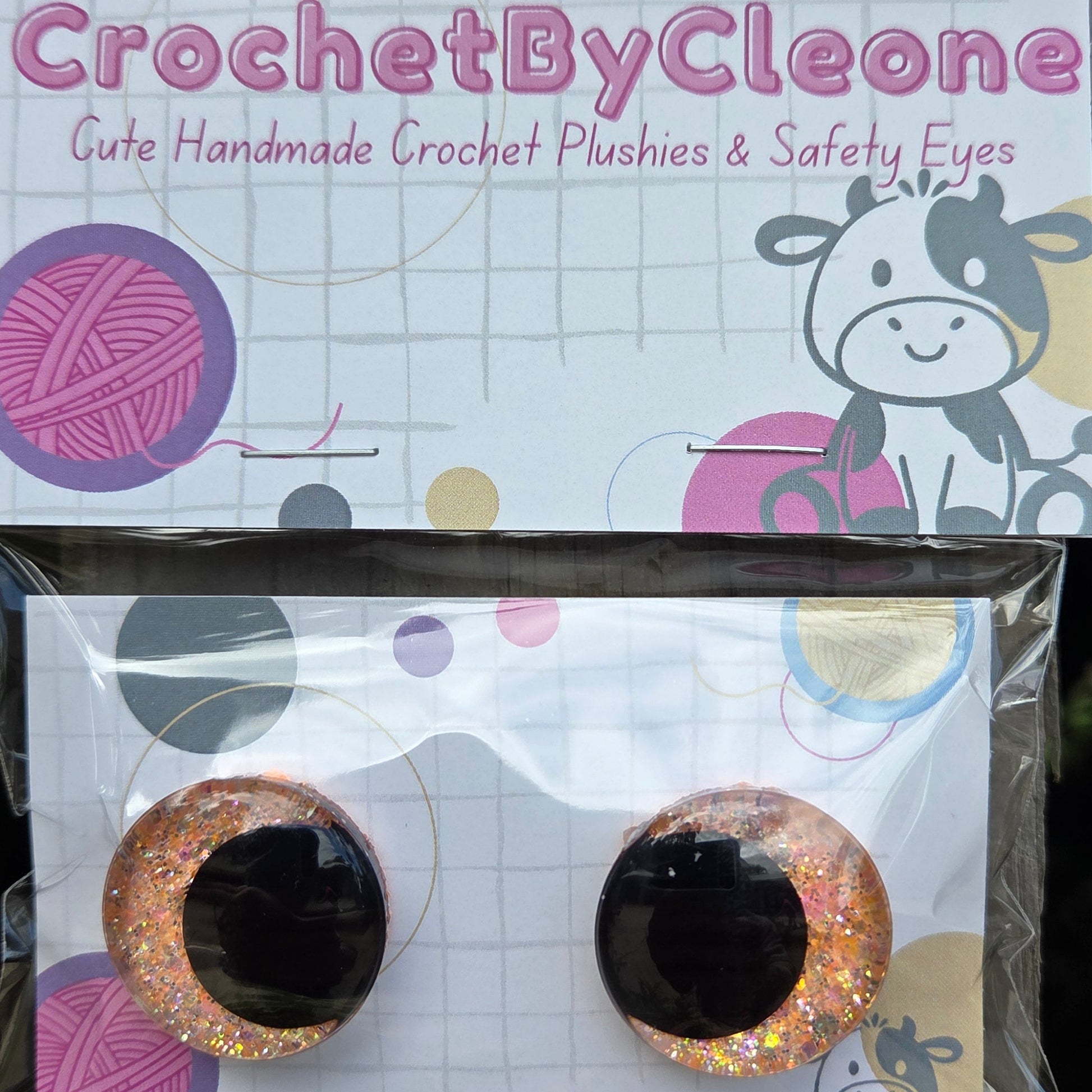 A pair of handmade crochet crochet eyes with glittery gold and pink edges and black centers, displayed on a branded card next to its packaging on a rustic wooden surface.