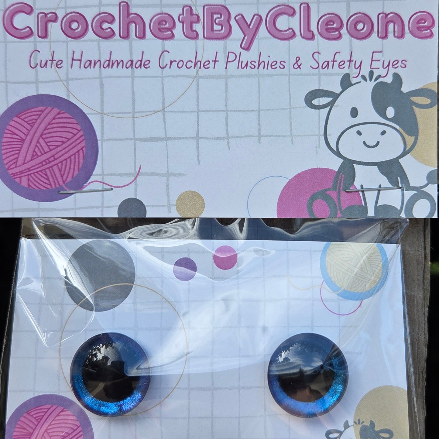 A packaged set of two blue and black crocheted safety eyes displayed on a branded backing card that reads 'CrochetByCleone' against a rustic wooden background.