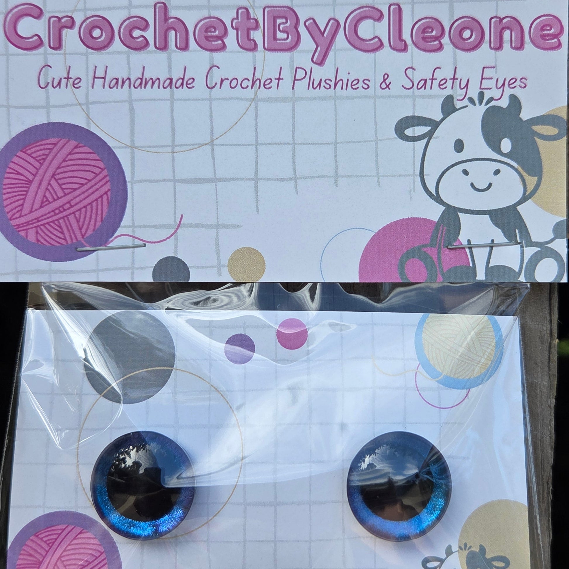 A packaged set of two blue and black crocheted safety eyes displayed on a branded backing card that reads 'CrochetByCleone' against a rustic wooden background.