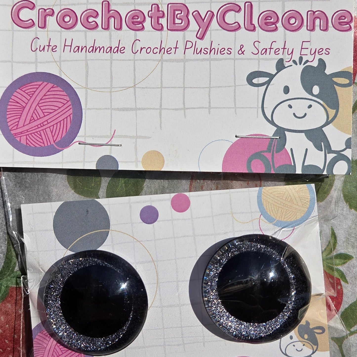 A pair of handmade crochet safety eyes with black glitter centers, mounted on a branded display card that reads 'CrochetByCleone' against a strawberry-patterned fabric background.