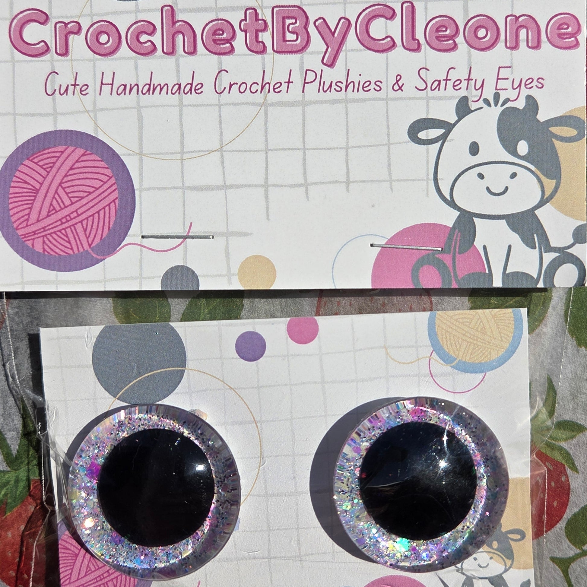 Two novelty crochet push eyes with glittery silver borders and black centers, displayed on branded white backing cards on a strawberry-patterned fabric surface.