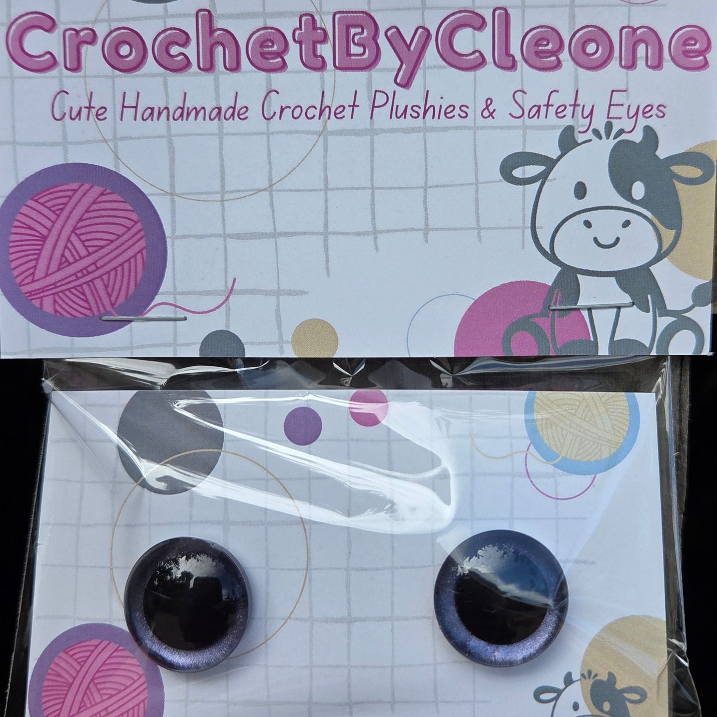 Crochet-style push-on safety eyes in a clear plastic sleeve next to their branded retail backing card. The product has two round glossy dark purple-blue eyes with black centers, mounted on a white card with crochet-themed graphics and the text 'CrochetByCleo'.