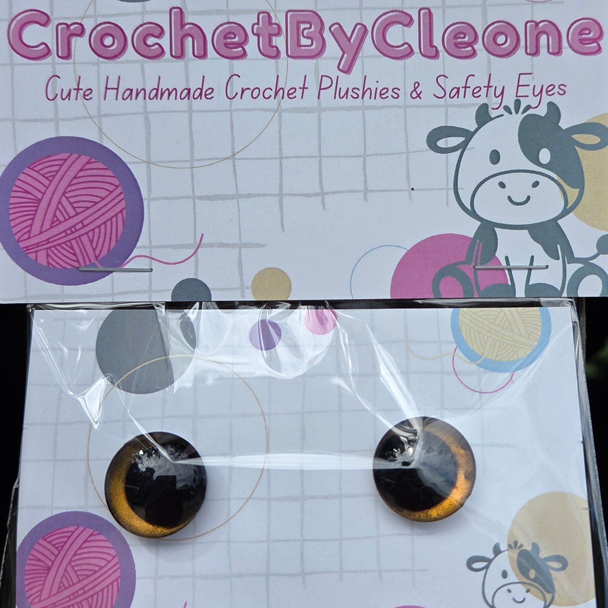 Two round crochet hook pushies with black and gold metallic ends, displayed on a branded white card inside a clear plastic sleeve, resting on a rustic wooden surface.