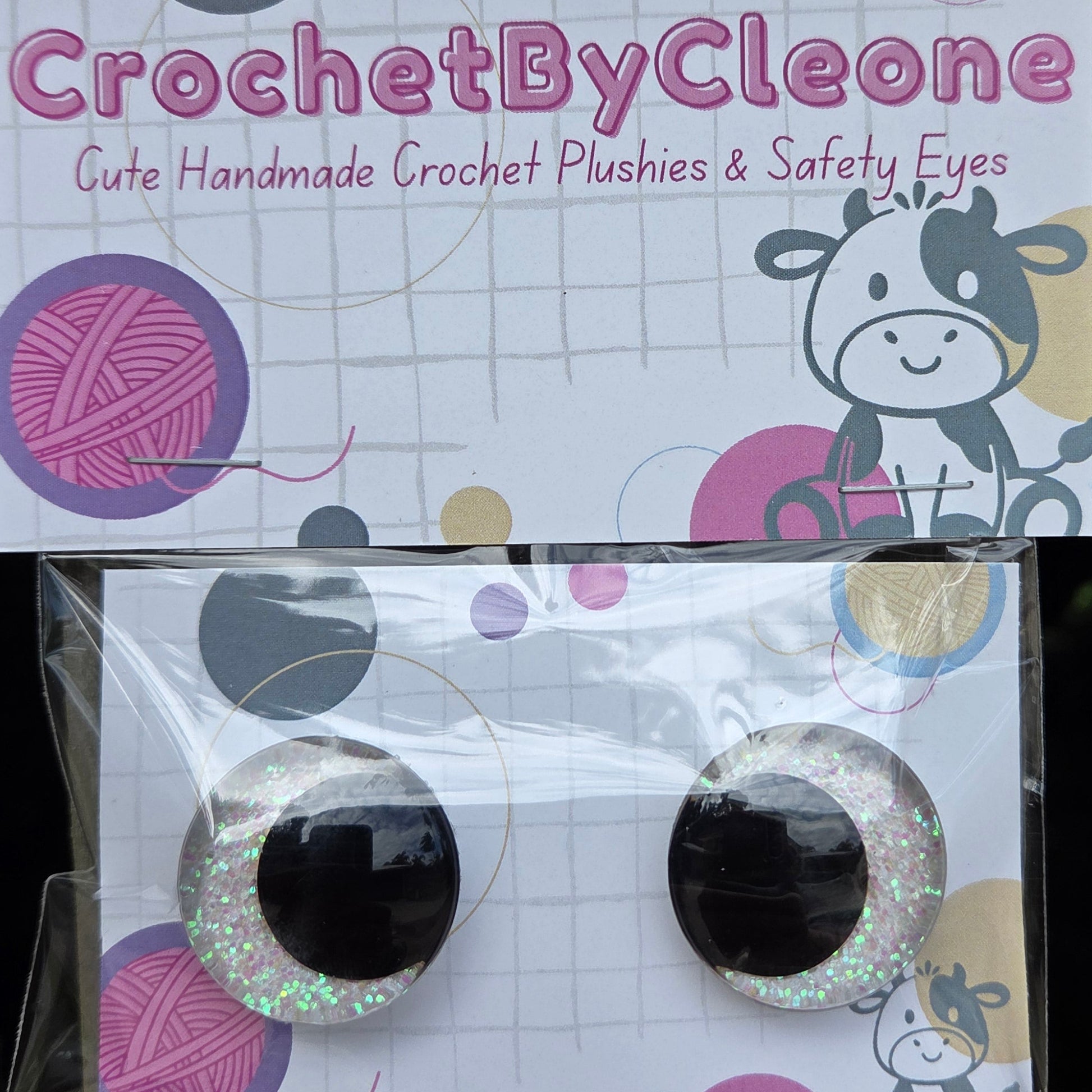 A pair of handmade crochet plushies and safety eyes displayed on branded backing cards, resting on a rustic wooden surface. The plushies feature black centers with one covered in silver glitter and the other in white glitter, alongside a pink and purple yarn ball motif.