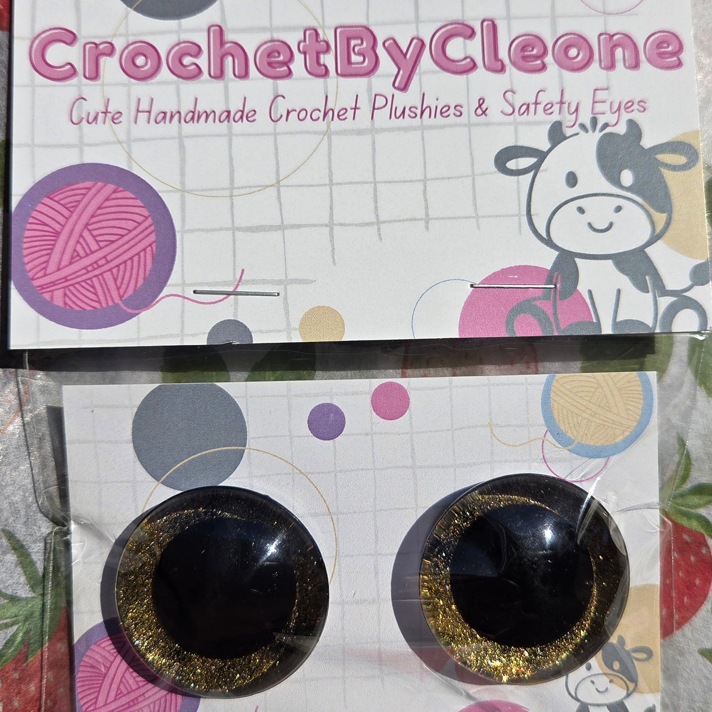 A pair of handmade crochet safety eyes with black centers and glittery gold edges, mounted on a branded display card for 'CrochetByCleone'.