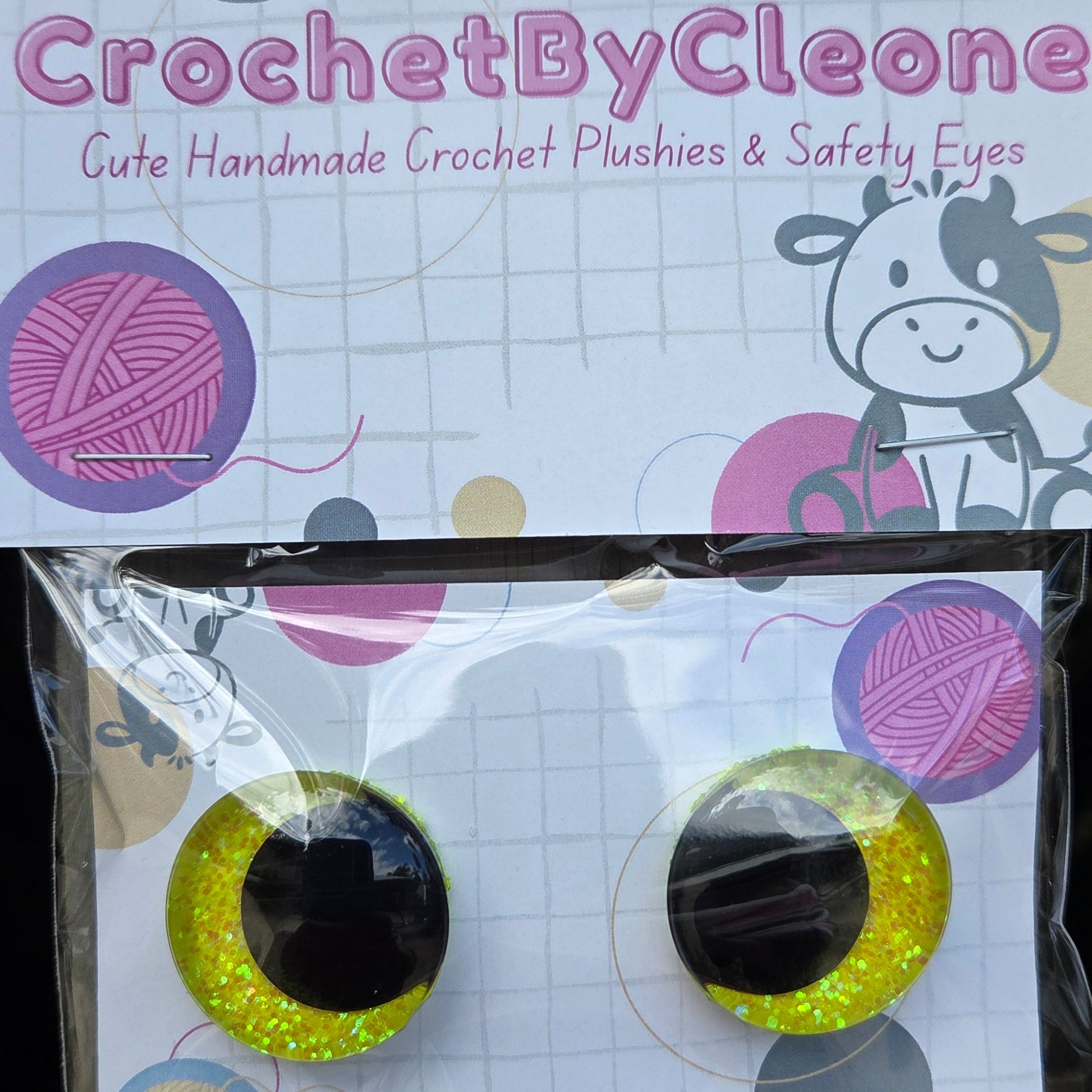 Two novelty crochet punch and safety eyes in glittery yellow and black, displayed on a white card with colorful yarn ball graphics. The card is branded 'CrochetByCleo' and placed on a rustic wooden surface next to its clear plastic packaging.