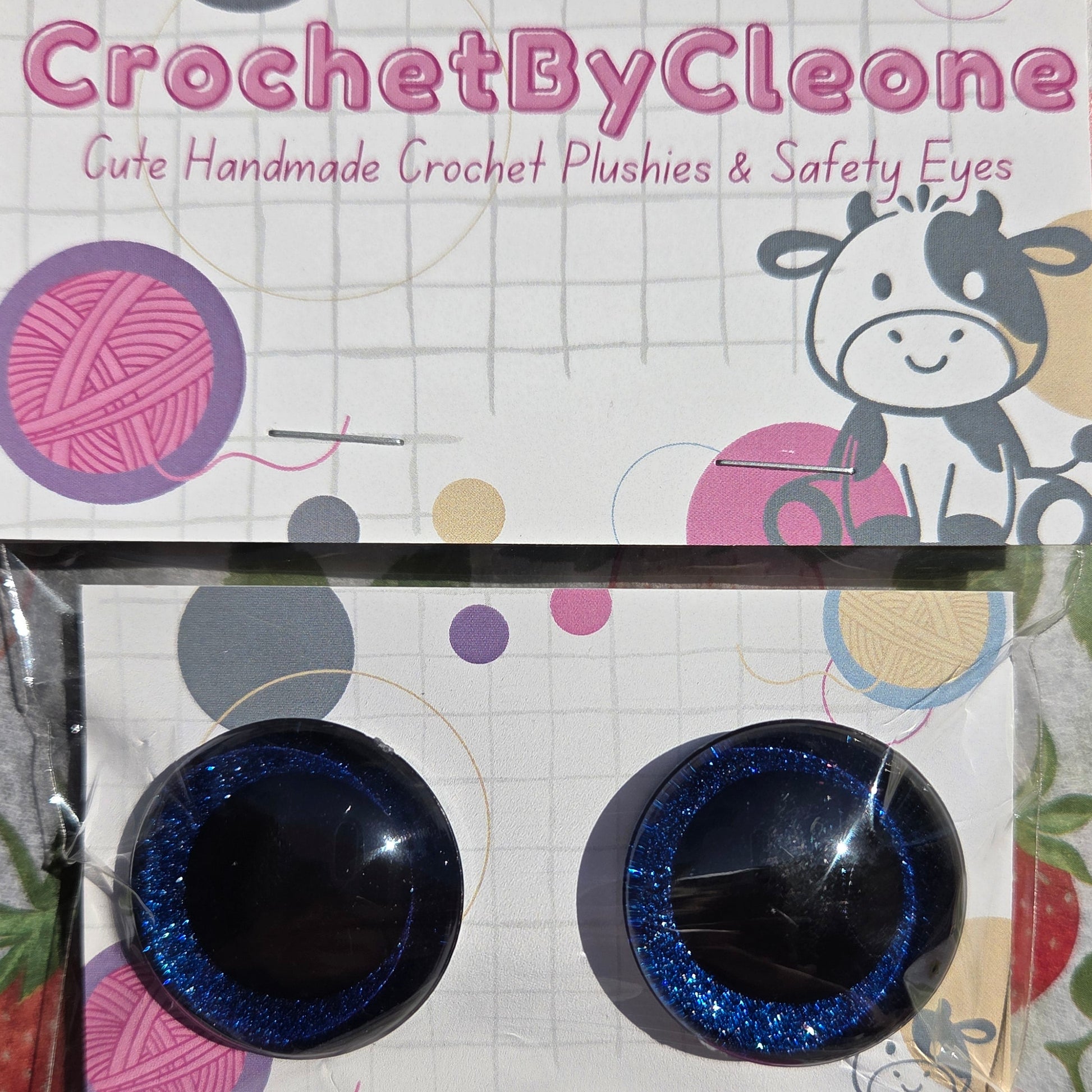 Two blue glittery safety eyes for crocheted dolls, shown with their printed packaging card on a strawberry-patterned fabric background. The brand 'CrochetByCleone' is visible on the packaging.
