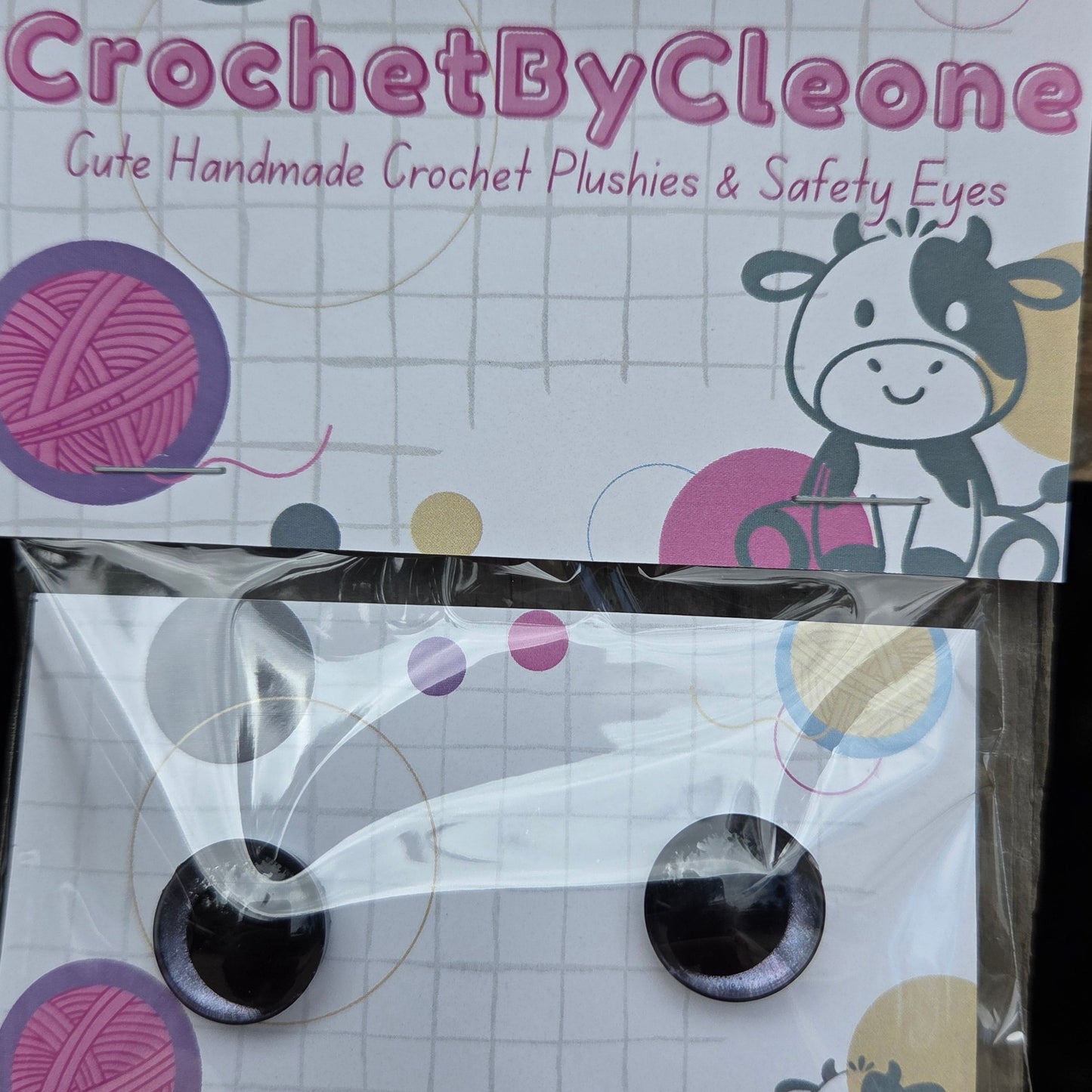 A pair of handmade crochet crochet safety eyes in a clear plastic sleeve, displayed next to its branded packaging card on a rustic wooden surface.