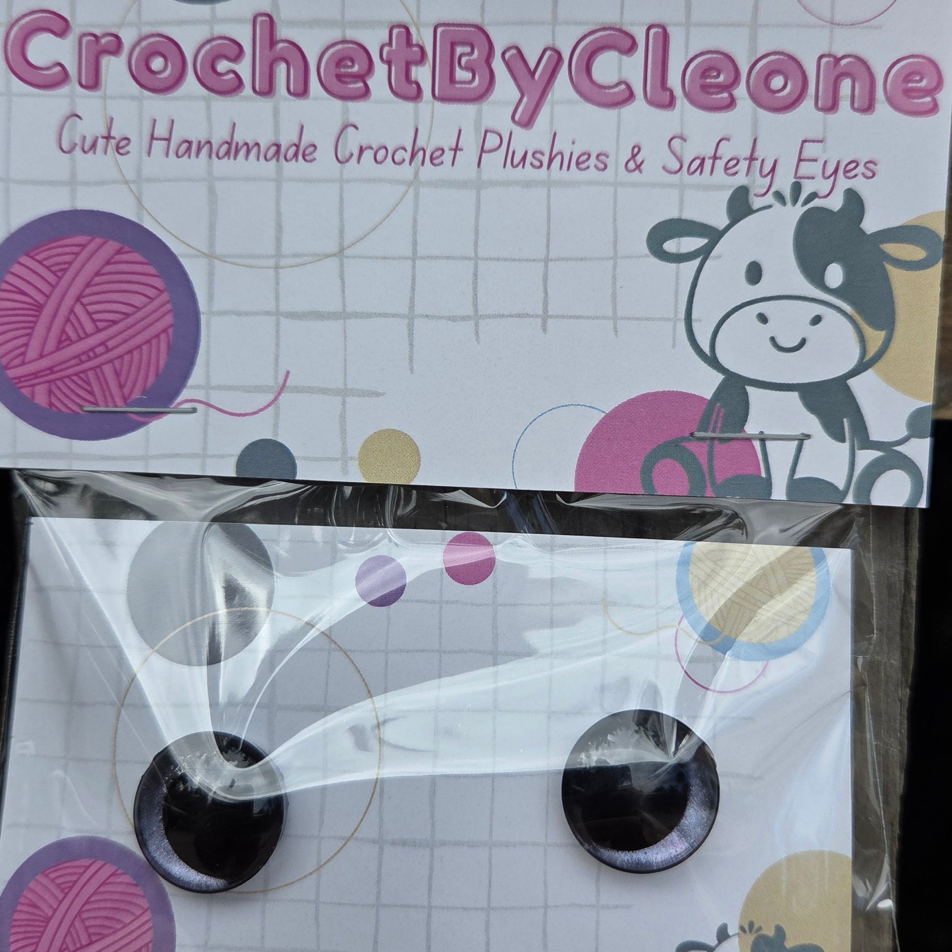 A pair of handmade crochet crochet safety eyes in a clear plastic sleeve, displayed next to its branded packaging card on a rustic wooden surface.