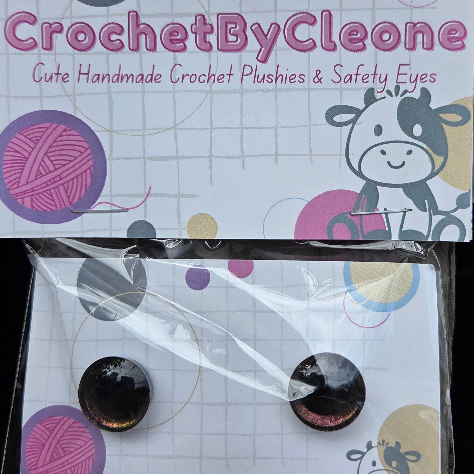CrochetByCleone handmade crochet plushies and safety eyes, shown in their retail packaging on a rustic wooden surface. The packaging features a white card with pink and purple yarn ball graphics and a cartoon cow.