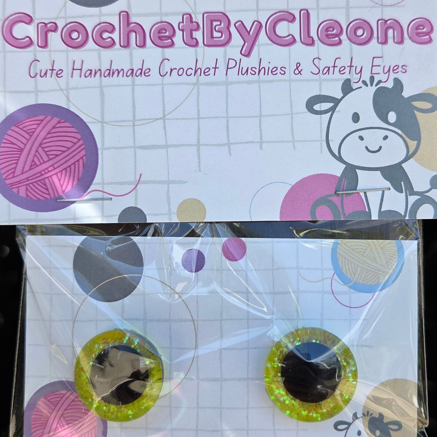 A pair of handmade crochet plushies with safety eyes, packaged in a clear plastic bag with a branded backing card. The card reads 'CrochetByCleone' and features colorful yarn ball graphics and a cartoon cow.