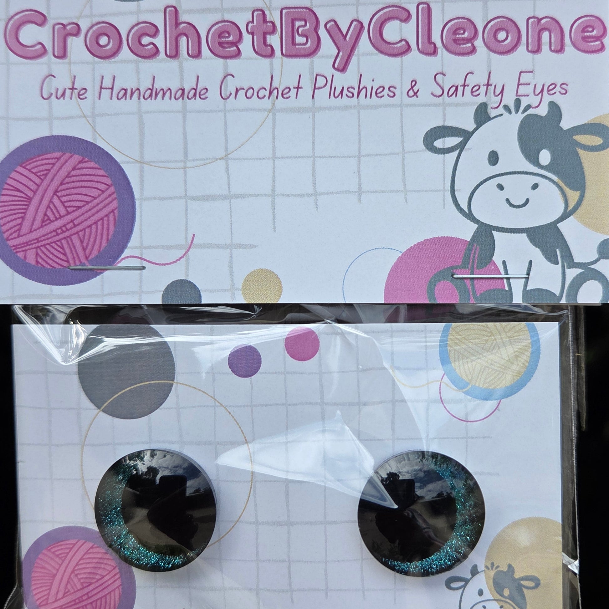 Two circular black crochet safety eyes with glittery teal-blue edges, displayed on a printed backing card next to the product packaging. The packaging features the brand name 'CrochetByCleo' and cartoon cow illustrations.