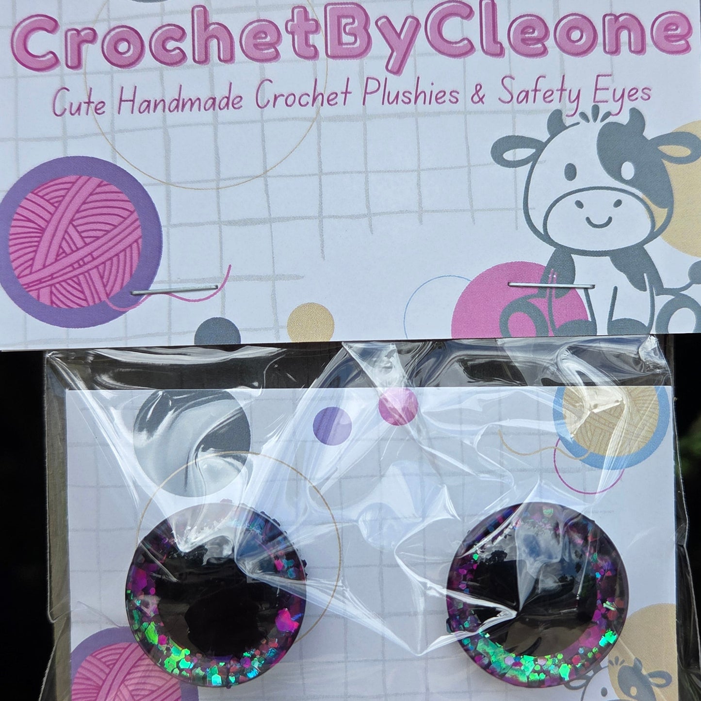 A pair of handmade crochet safety eyes in a clear plastic bag with a branded backing card, placed on a rustic wooden surface. The brand 'CrochetByCleone' is clearly visible on the card.