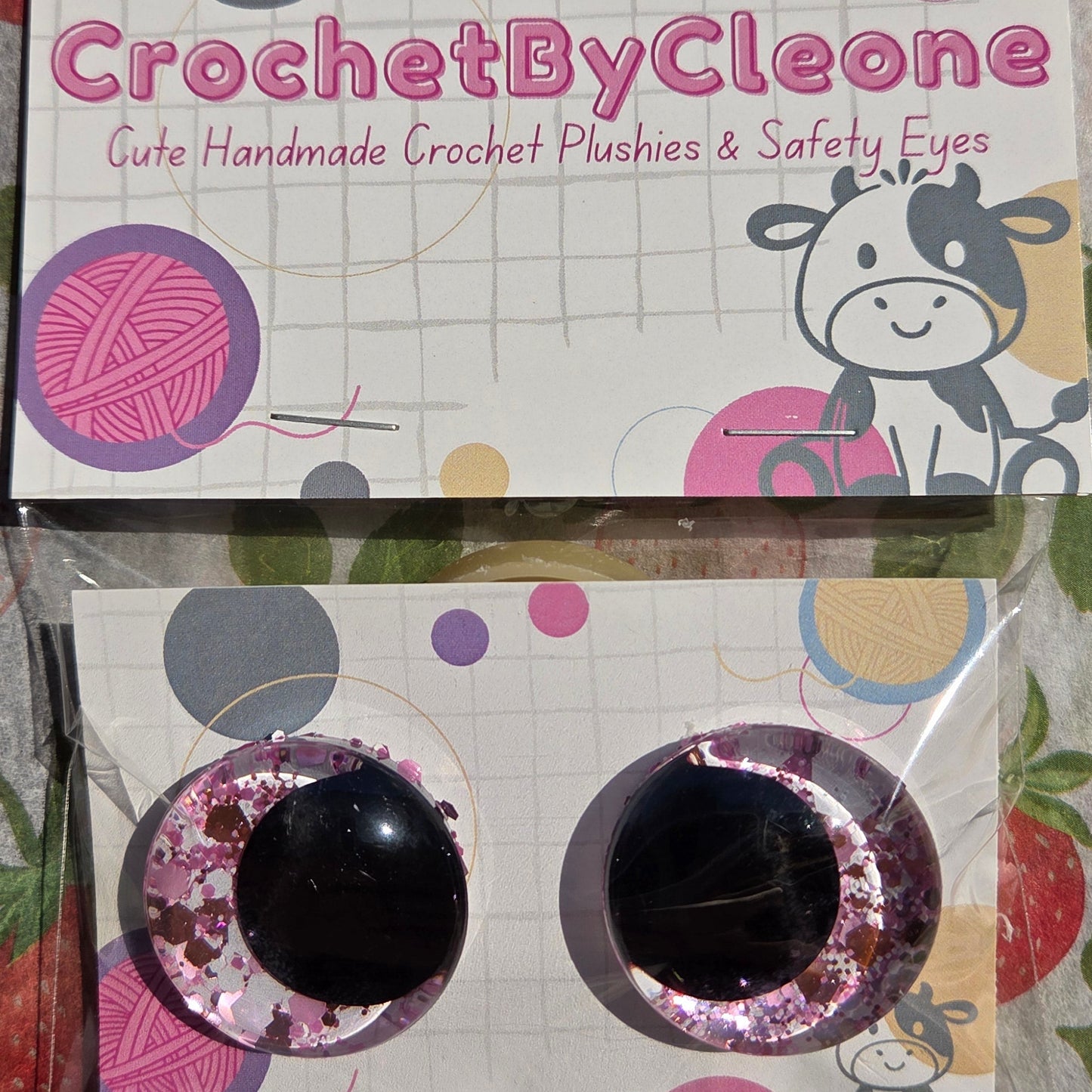 Two packaged crochet hook sets displayed on printed cards over a strawberry-patterned fabric surface. The left package is labeled 'CrochetByCleo' and the right package contains two black crochet hooks with pink glitter trim.