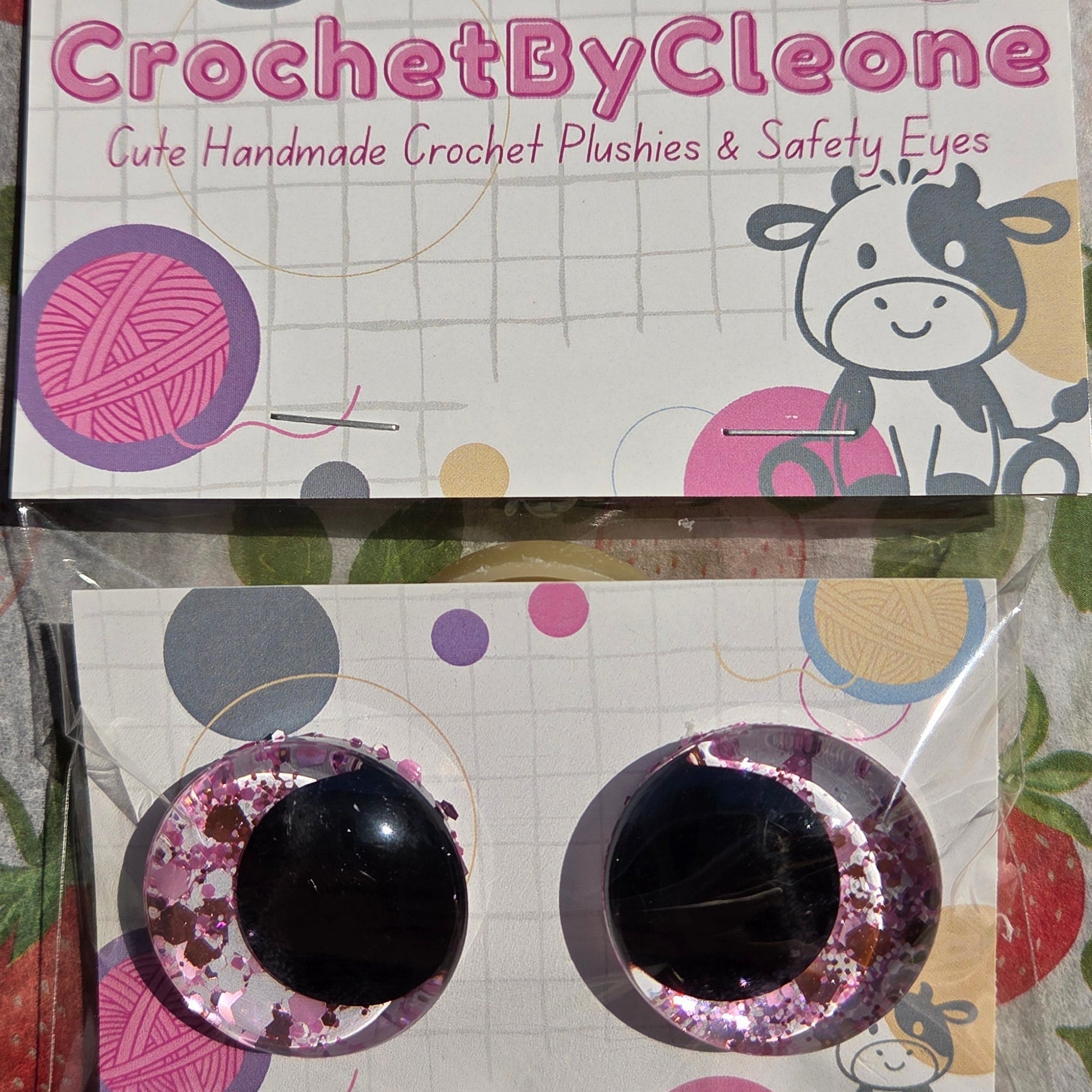 Two packaged crochet hook sets displayed on printed cards over a strawberry-patterned fabric surface. The left package is labeled 'CrochetByCleo' and the right package contains two black crochet hooks with pink glitter trim.