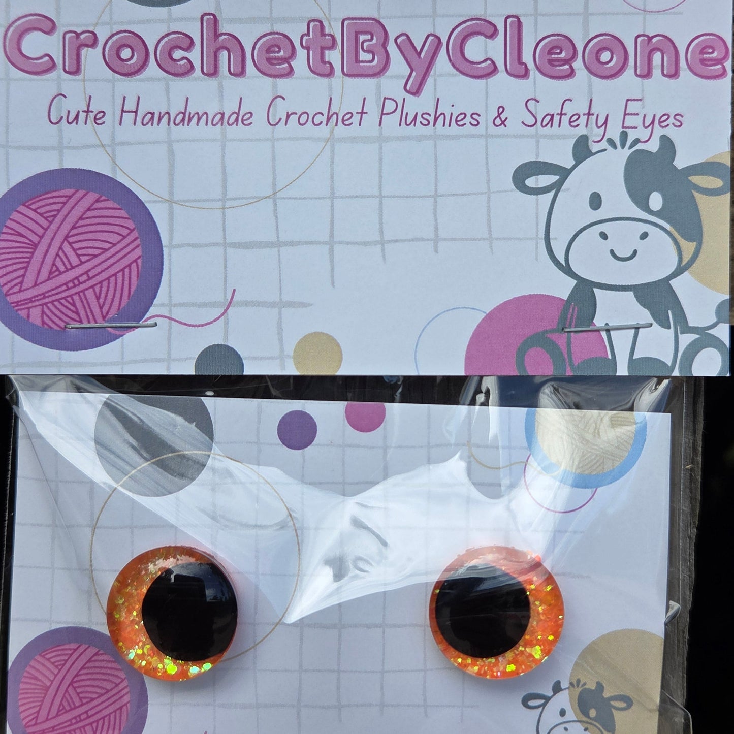 A pair of handmade crochet eye embellishments in clear plastic packaging with a branded backing card. The product consists of two round, glittery orange-and-black crochet eyes, and the backing card features the brand name 'CrochetByCleone' and decorative yarn ball graphics.