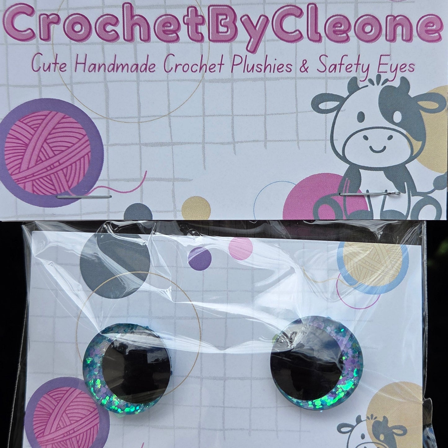 A pair of handmade crochet push-on safety eyes in a clear plastic sleeve, displayed next to its printed packaging card on a rustic wooden surface. The packaging features pink yarn ball graphics and the brand name 'CrochetByCleo'.