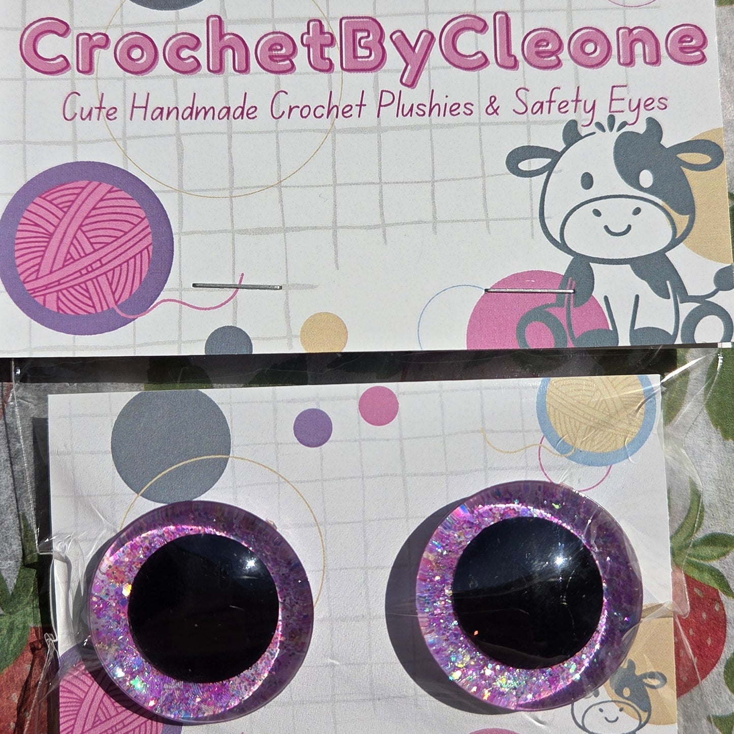 A pair of handmade crochet safety eyes with glittery purple and pink borders and black centers, attached to a branded display card for 'CrochetByCleone'.