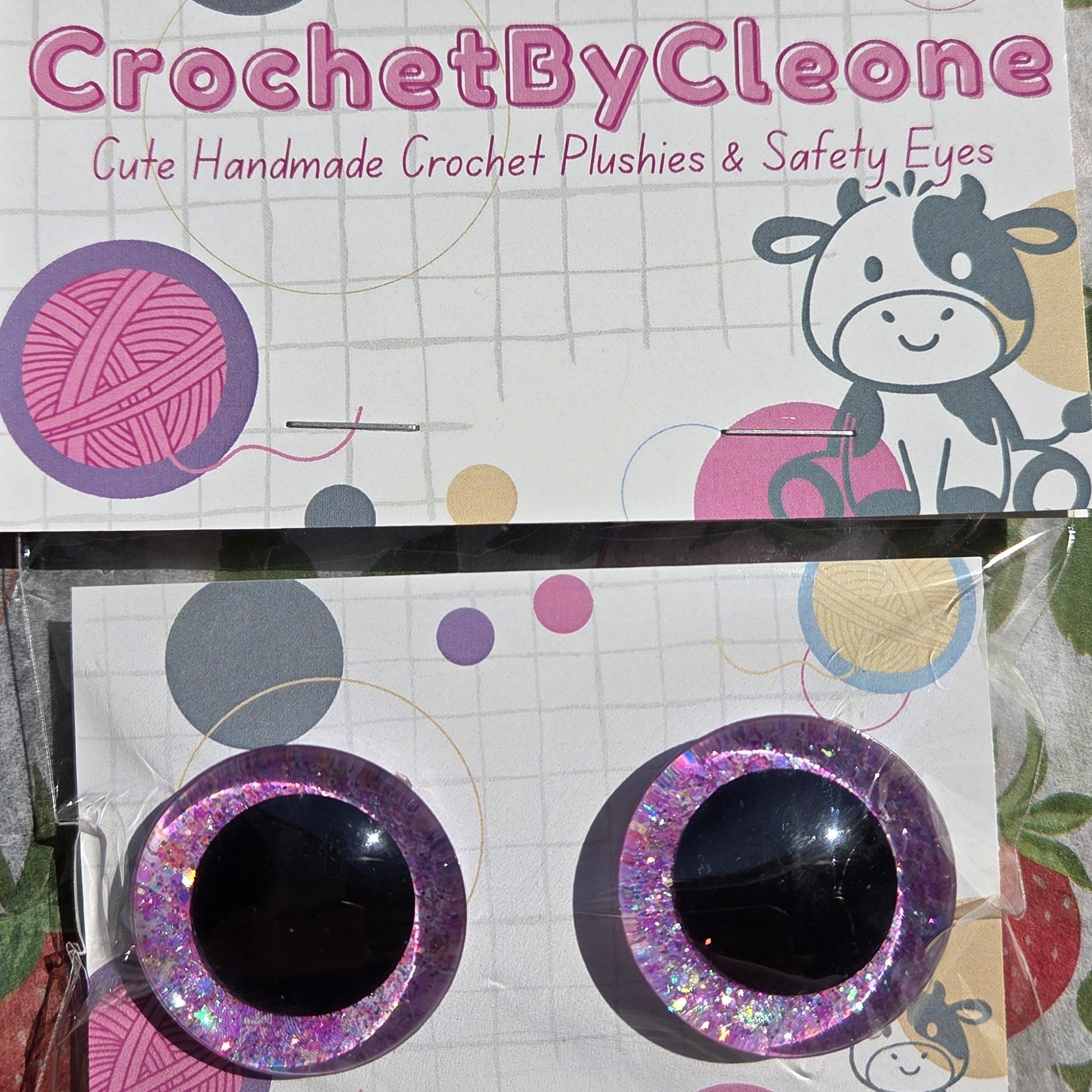 A pair of handmade crochet safety eyes with glittery purple and pink borders and black centers, attached to a branded display card for 'CrochetByCleone'.