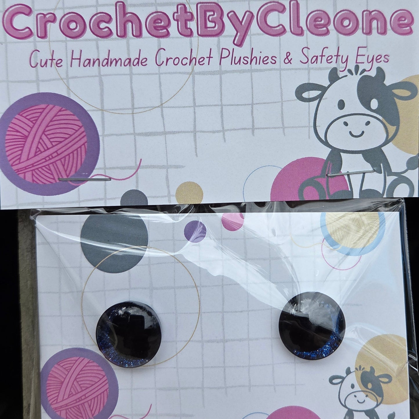 Two circular crochet hook safety eyes with black centers and blue outer rings, displayed on a white printed backing card and a clear plastic sleeve, set on a rustic wooden surface.