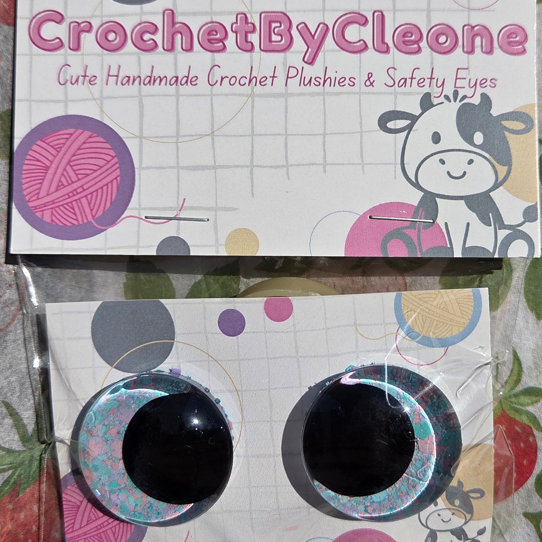 Crochet starter kit contents displayed on a strawberry-patterned fabric background, including a printed package labeled 'CrochetByCleo' and a small plastic bag containing two black crochet hooks.