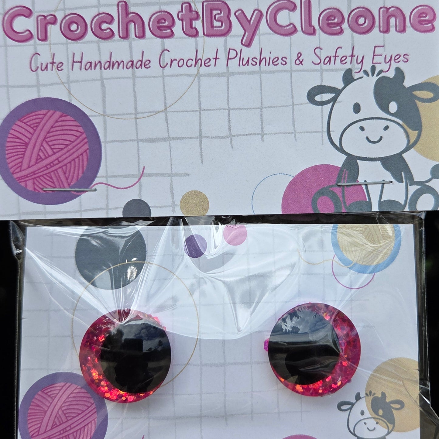 A pair of round pink glittery crochet crochet hooks in their retail packaging, displayed next to the product card on a rustic wooden surface.