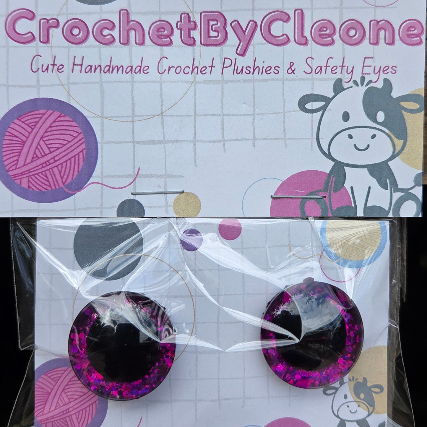 A pair of handmade crochet plastic and safety eyes in a purple glittery design, packaged on a white card with a cow illustration and the brand name 'CrochetByCleo'.