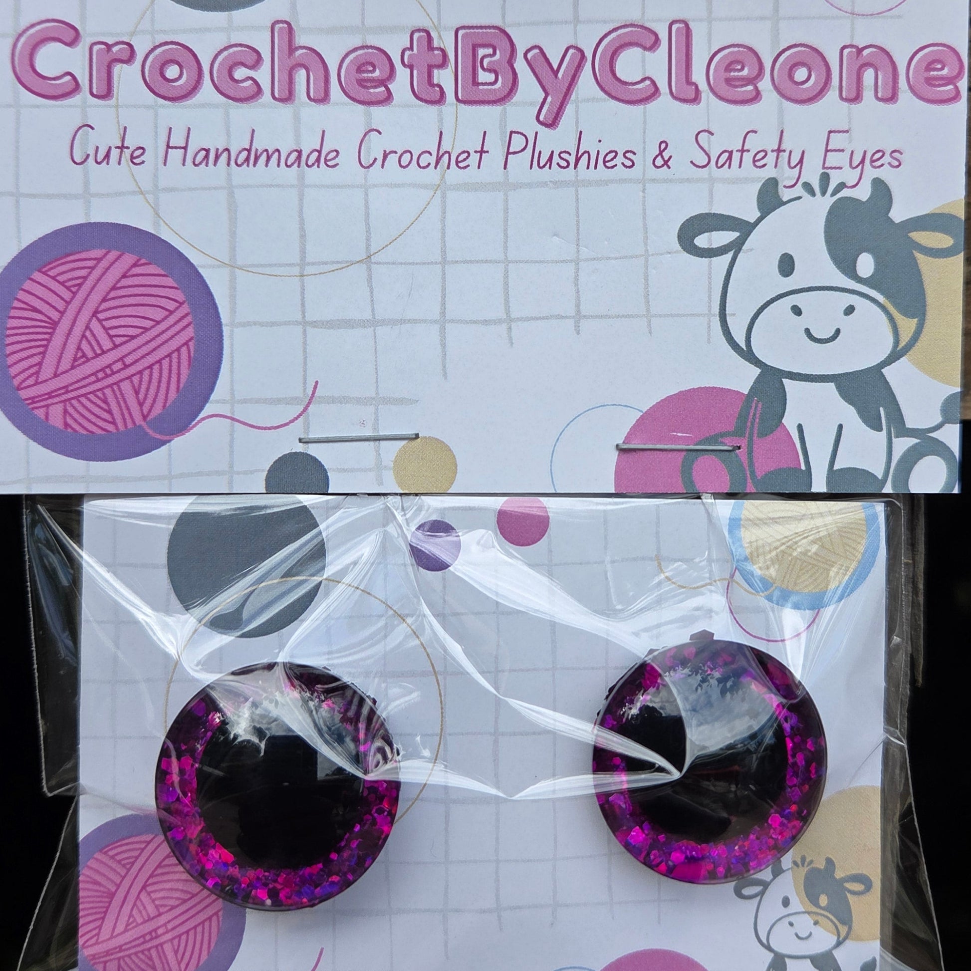 A pair of handmade crochet plastic and safety eyes in a purple glittery design, packaged on a white card with a cow illustration and the brand name 'CrochetByCleo'.