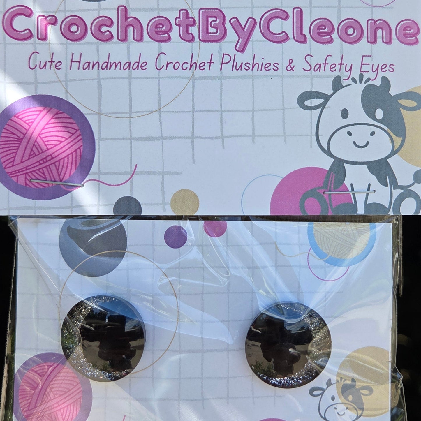 Two packaged crochet hooks with decorative printed cards on a rustic wooden surface. The left card reads 'CrochetByCleo' and the right package shows the hooks through clear plastic with matching circular packaging.