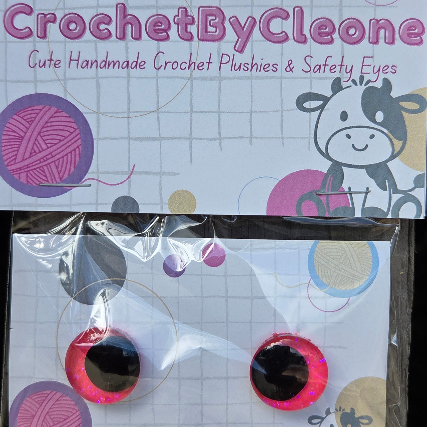 Packaged crochet safety eyes displayed on a branded card. The card reads 'CrochetByCleone' and shows the product in a clear plastic bag with two round pink-and-black safety eyes attached to a printed white backing card featuring yarn balls, circles, and a cow illustration.