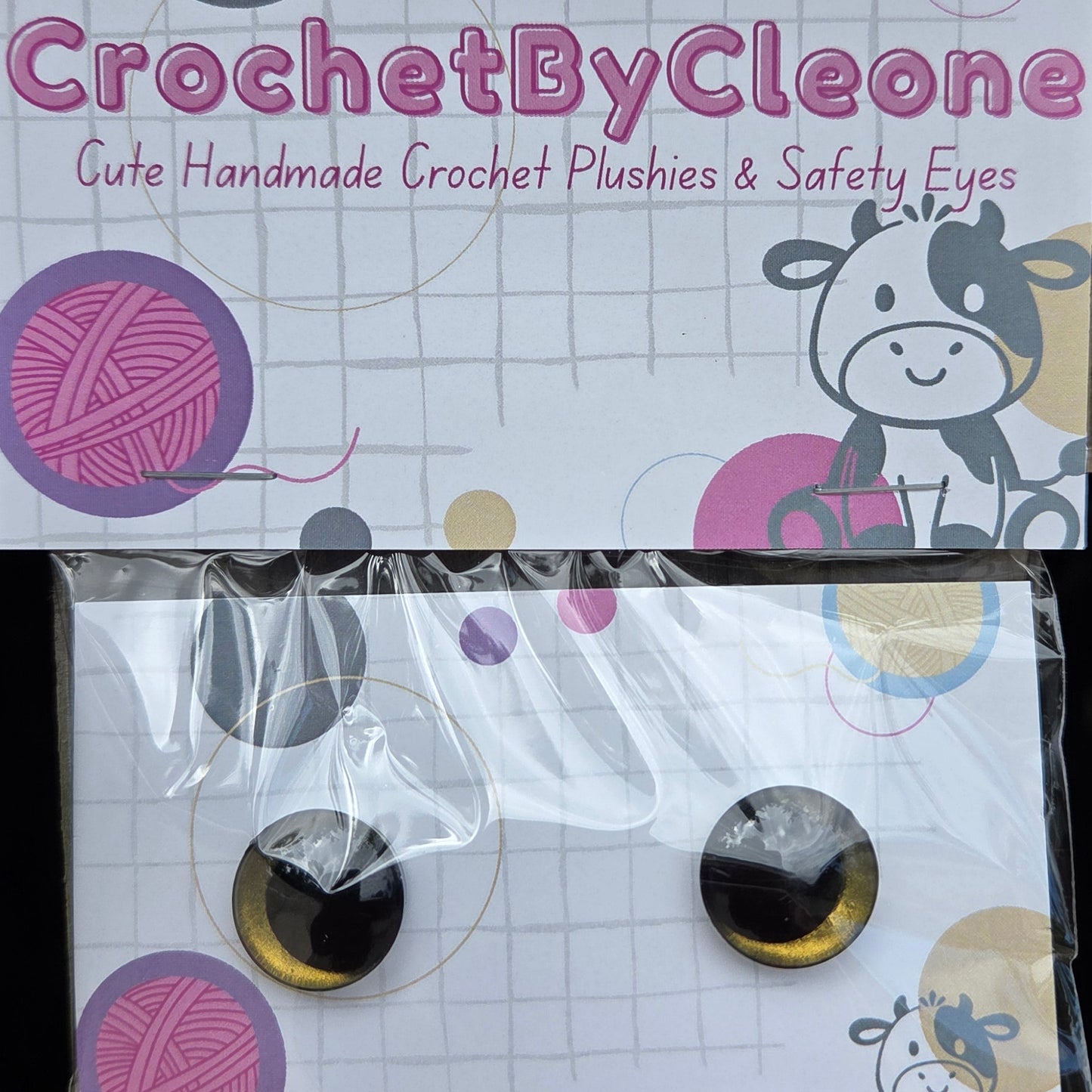 Two crochet plush eyes with black and gold reflective pupils, displayed on branded white backing cards on a rustic wooden surface. The left card shows the brand name 'CrochetByCleo' and the right card shows the packaged pair of eyes.