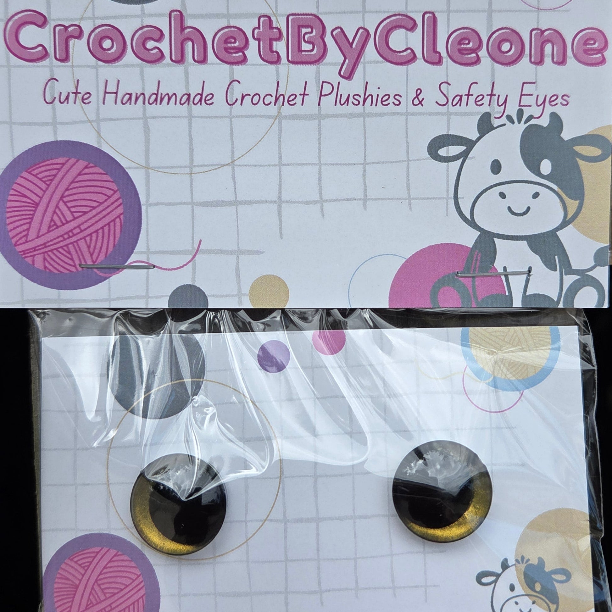 Two crochet plush eyes with black and gold reflective pupils, displayed on branded white backing cards on a rustic wooden surface. The left card shows the brand name 'CrochetByCleo' and the right card shows the packaged pair of eyes.