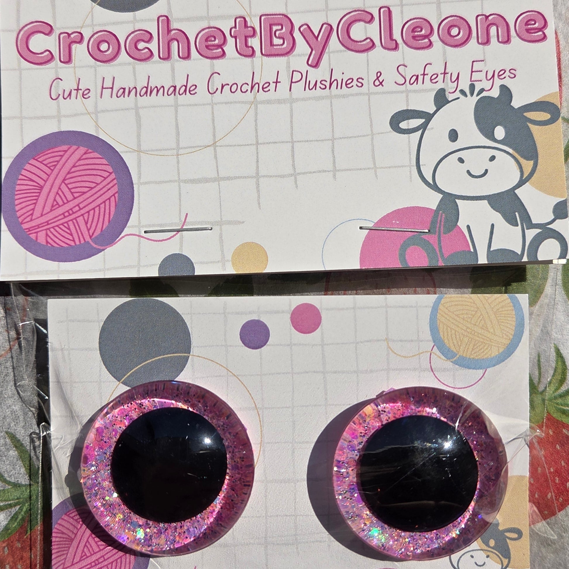 Two novelty crochet hook holders or cards displayed on a strawberry-patterned fabric background. The left card is white with pink and gray yarn-themed graphics and the vertical text 'CrochetByCleo'.