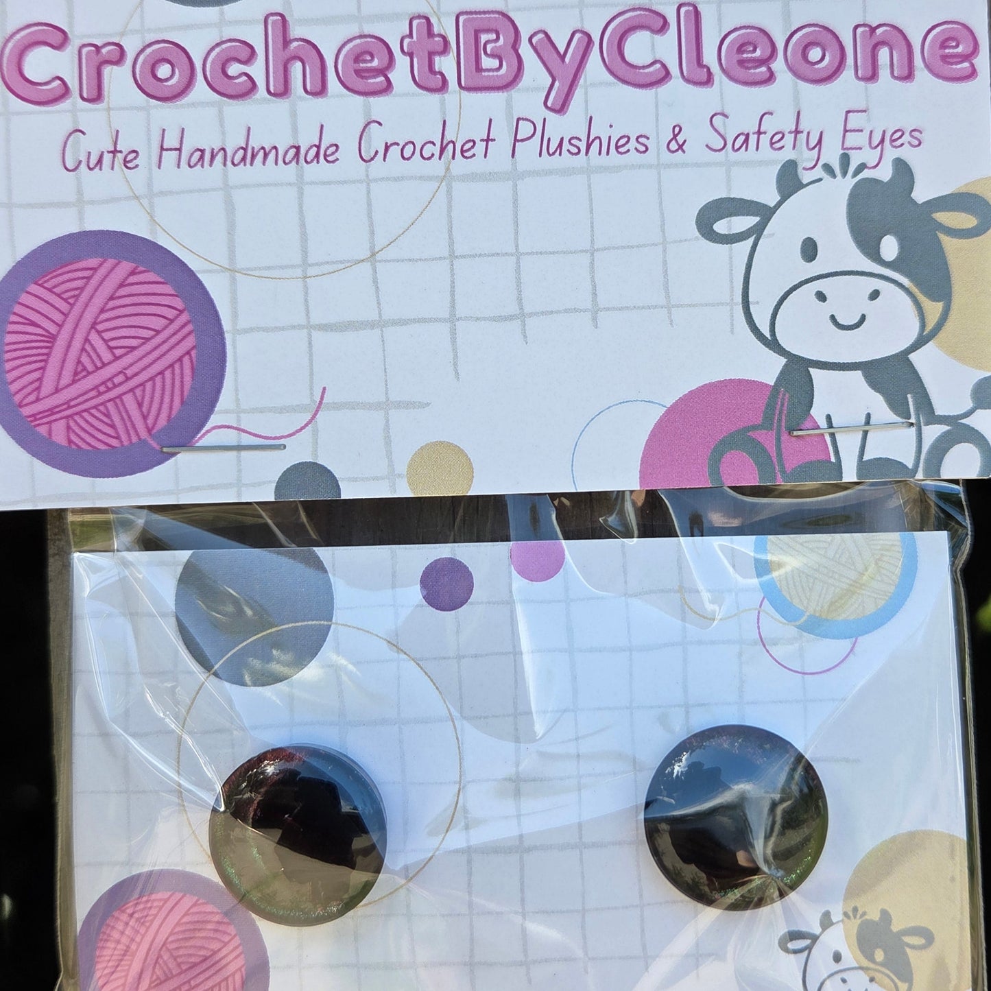 A pair of handmade crochet plugs and safety eyes in circular shapes, packaged on a printed card labeled 'CrochetByCleo' alongside a matching card insert, photographed on a rustic wooden surface.