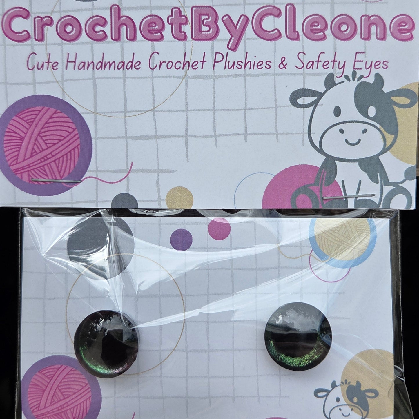 A pair of handmade crochet plushies in clear plastic packaging with a branded backing card. The plushies are round and have black centers with iridescent green outer edges.